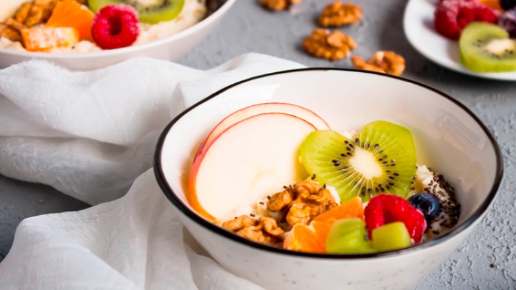 Verschiedene Schalen mit Haferbrei, dekoriert mit Kiwi, Apfelscheiben, Beeren, Mandarinenstücken und Walnüssen auf grauem Hintergrund.