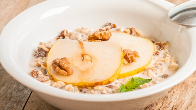 Weiße Schale mit Müsli in Milch, dekoriert mit zwei Birnenscheiben, Walnüssen und einem Minzblatt, auf Holzuntergrund.