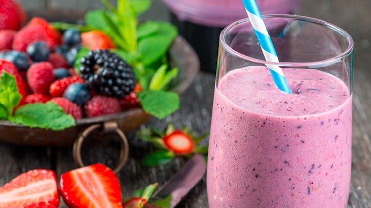 Ein Glas lila Beerensmoothie mit blau-weißem Strohhalm steht neben frischen Beeren, Minzblättern und einem Messer auf Holzuntergrund.