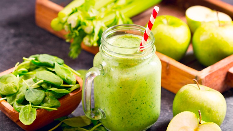 Ein grüner Smoothie im Glas mit rotem Strohhalm, umgeben von frischem Spinat, Stangensellerie und grünen Äpfeln.