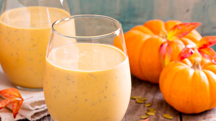 Zwei Gläser mit orangefarbenem Kürbis-Smoothie stehen auf einer Holzfläche, daneben kleine Kürbisse, rote Blätter und Kürbiskerne.
