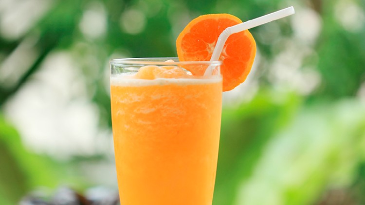 Orangener Fruchtshake in einem Glas mit weißem Trinkhalm, dekoriert mit einer halben Orangenscheibe, auf Bambusmatte.