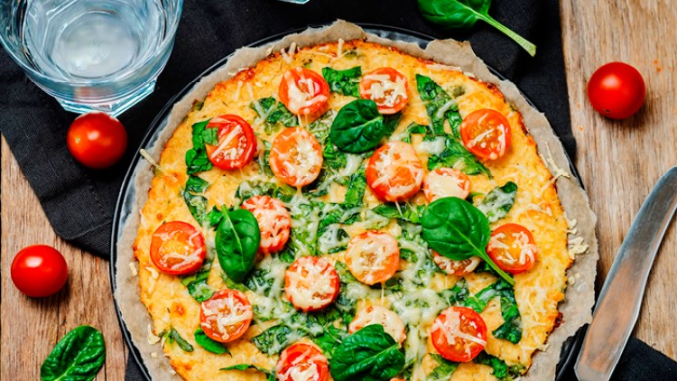 Knusprige Pizza mit Basilikum, Kirschtomaten, Käse und Spinatblättern liegt auf Backpapier, daneben Messer, Gabel und Trinkglas.