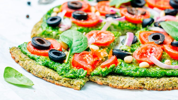 Grüne Pizza mit Spinatboden, Pesto, Tomatenscheiben, schwarzen Oliven, Zwiebeln, Pinienkernen und frischen Spinatblättern.