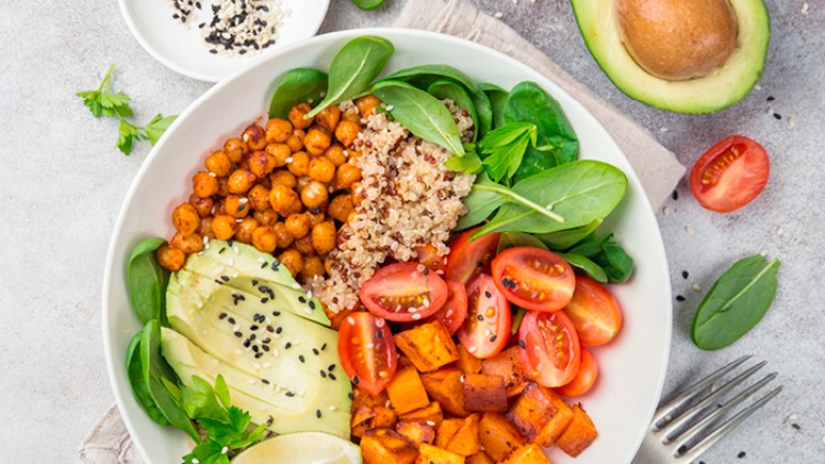 Eine weiße Schale mit Avocado, Kichererbsen, Quinoa, Süßkartoffeln, Tomaten und Blattspinat, garniert mit Sesam und Limette.