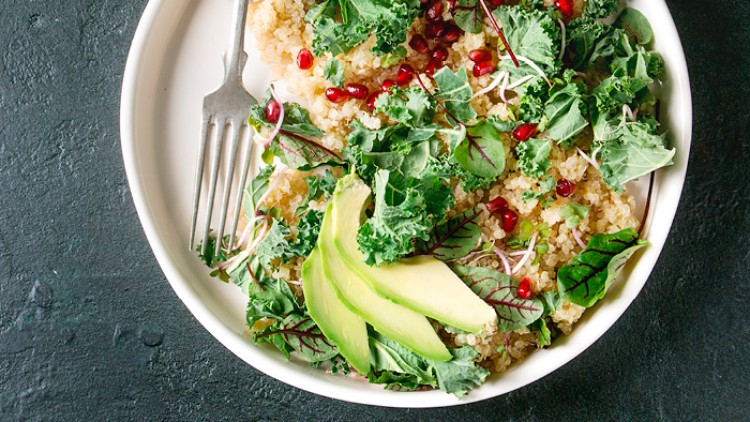 Schale mit Quinoa, Grünkohl, Avocadoscheiben, Granatapfelkernen und gemischtem Blattgrün, daneben eine Gabel.