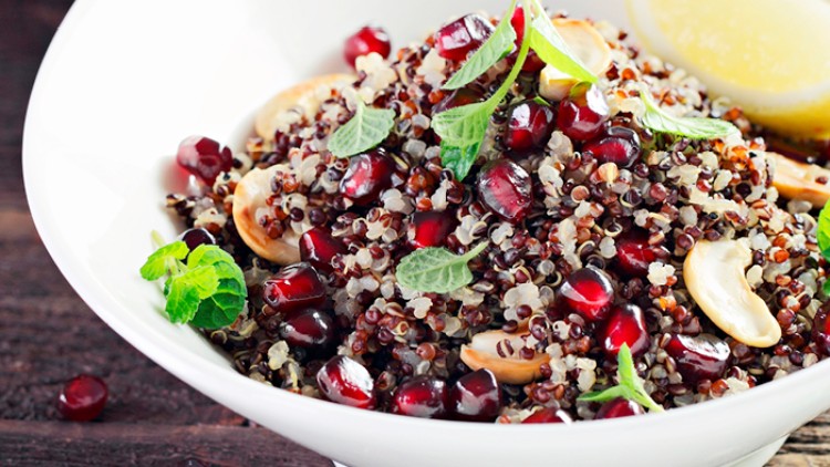 Schale mit Quinoa, Granatapfelkernen, Cashewkernen, frischer Minze und einer Zitronenspalte auf dunklem Untergrund.