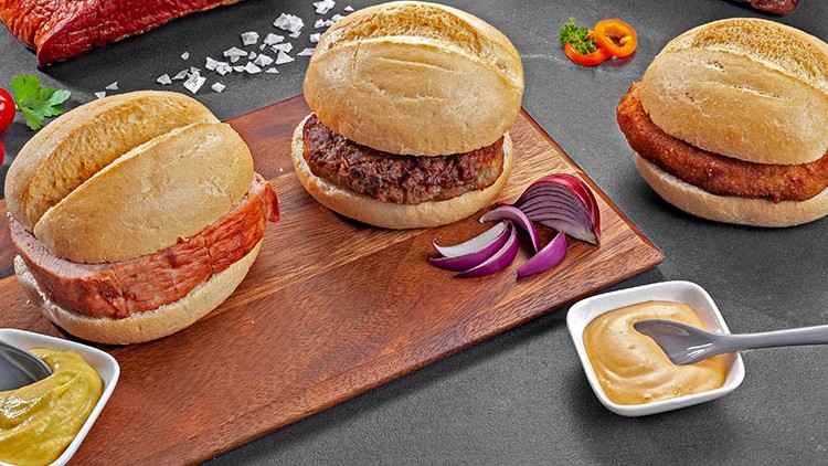 Drei belegte Brötchen mit Leberkäse, Frikadelle und Schnitzel auf Holzbrett, dazu Senf, Soße, Cherrytomaten und Zwiebeln.