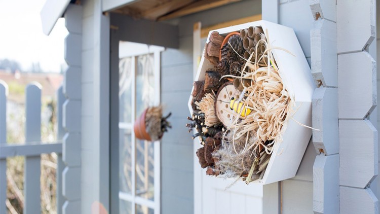 Ein sechseckiges Insektenhotel mit Naturmaterialien hängt an einer grauen Gartenhauswand neben einem Blumenübertopf.