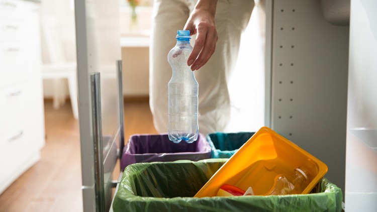 Eine Hand wirft eine leere Plastikflasche in einen offenen Abfallbehälter mit unterschiedlich farbigen Müllsortierfächern.