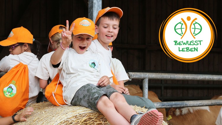 Kinder in weißen T-Shirts und orangefarbenen Kappen sitzen auf Strohballen, daneben das Logo „BEWUSST LEBEN“.