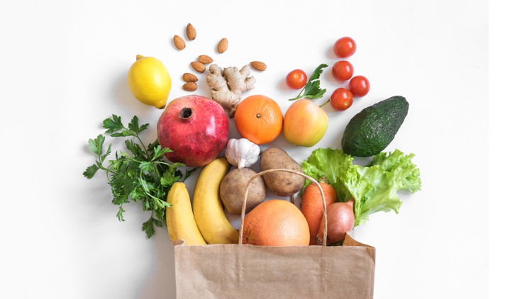 Eine gefüllte braune Papiertüte mit Obst, Gemüse und Nüssen, darunter Banane, Avocado, Granatapfel und Kopfsalat auf weißem Hintergrund.