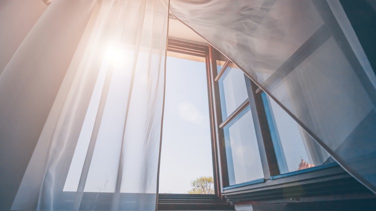 Geöffnetes Fenster mit weißem, transparentem Vorhang, durch das Sonnenlicht und ein blauer Himmel mit wenigen Details scheint.
