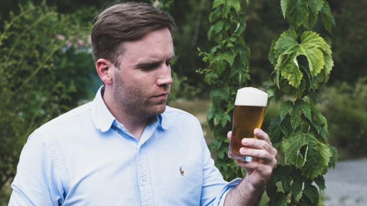 Person in blauem Hemd hält ein Glas Bier mit Schaumkrone in der Hand, im Hintergrund grüne Hopfenpflanzen.