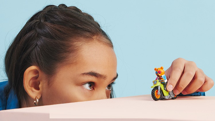 Eine Hand hält ein kleines grünes Spielzeug-Motorrad, auf dem eine bunte Tierfigur sitzt, vor einem blauen Hintergrund.