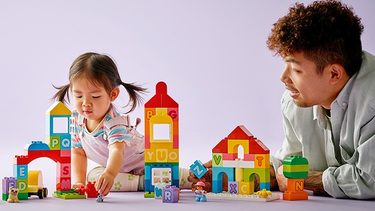 Ein Kind und eine weitere Person spielen mit bunten Bauklötzen und Figuren in einem hellen Raum auf einem Tisch.