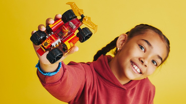 Eine Hand hält ein rotes Spielzeugauto mit großen schwarzen Rädern und goldfarbenem Heckflügel vor gelbem Hintergrund.