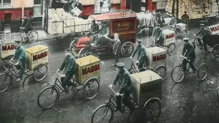 Männer in Uniform fahren mit Fahrrädern, die gelbe Kisten mit rotem MAGGI-Schriftzug transportieren, durch eine Straße.