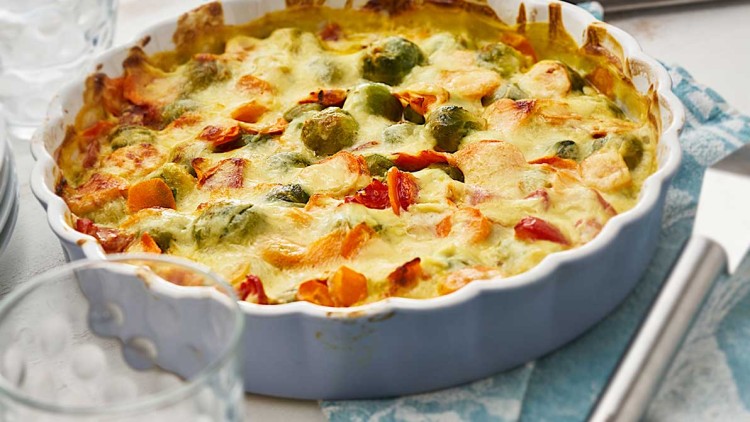 Gratin mit überbackenem Gemüse wie Rosenkohl, Karotten und Paprika in einer weißen Auflaufform auf einem blauen Tuch.