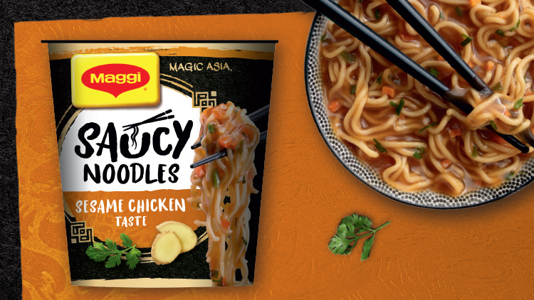 Maggi-Becher mit der Aufschrift „Saucy Noodles Sesame Chicken Taste“ neben einem Teller mit Nudeln und schwarzen Essstäbchen.