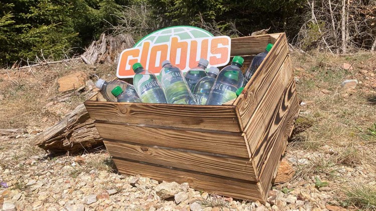 Holzkiste mit mehreren Mineralwasserflaschen und GLOBUS-Logo vor einem Hintergrund aus Wald, Wiese und Kiesboden.