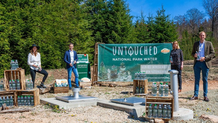 Ein Banner mit der Aufschrift „UNTOUCHED NATIONAL PARK WATER“ steht vor Bäumen, daneben Kisten mit Mineralwasserflaschen.