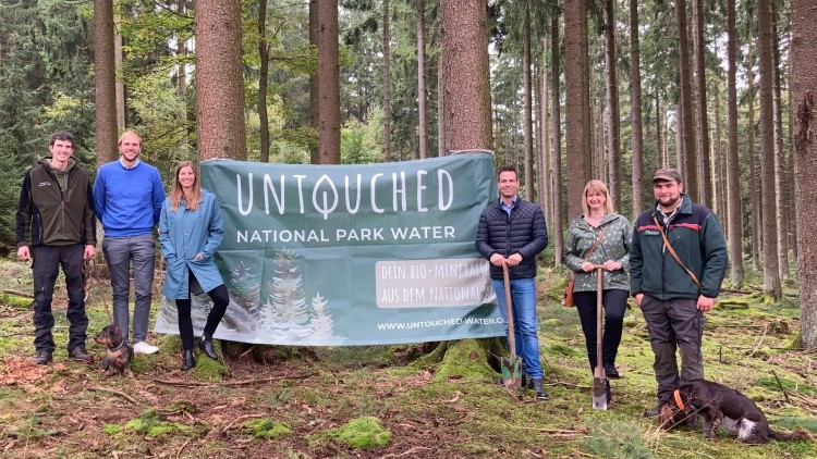 Sechs Personen stehen mit zwei Hunden vor einem Banner im Wald, darauf steht: \