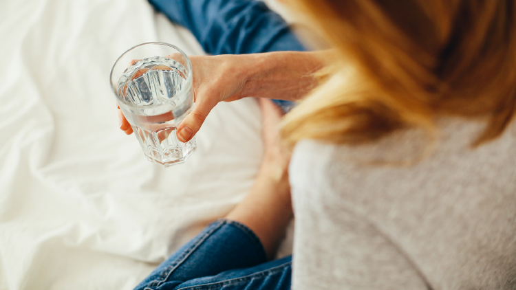 Eine Person hält in der rechten Hand ein Glas Wasser, im Hintergrund Bettwäsche und blaue Kleidung erkennbar.