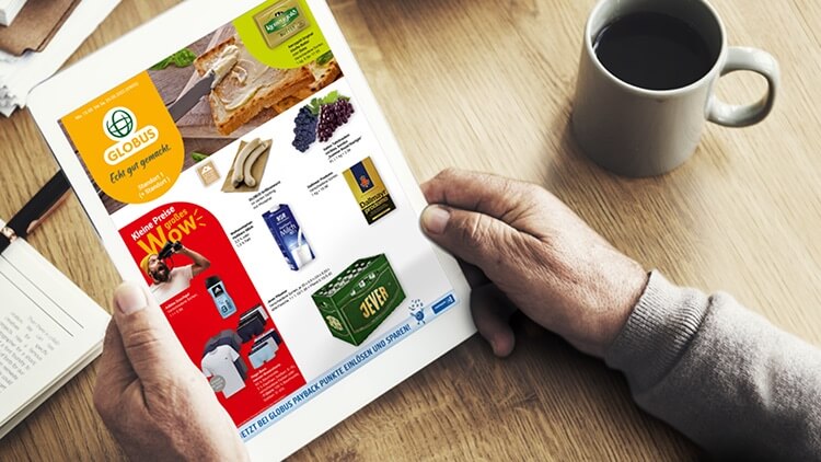 Eine Person hält ein Tablet mit einer GLOBUS-Werbeanzeige, daneben stehen eine Tasse Kaffee und ein Notizbuch auf einem Holztisch.