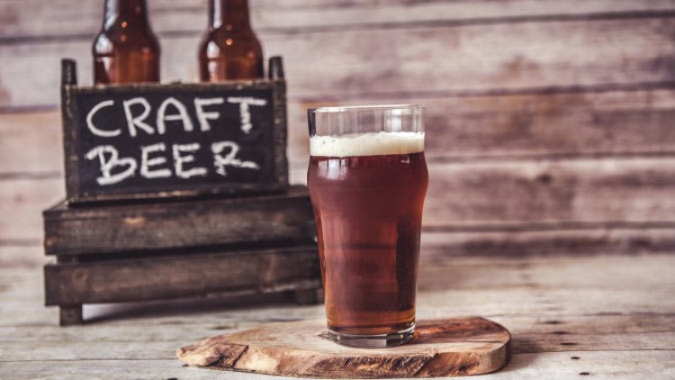 Ein Glas mit bernsteinfarbenem Craft-Bier steht auf einem Holzbrett, im Hintergrund eine Holzkiste mit der Aufschrift Craft Beer.