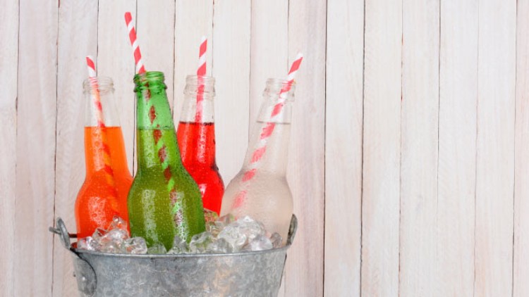 Vier Glasflaschen mit bunten Getränken und gestreiften Strohhalmen stehen in einem Eimer mit Eiswürfeln vor einer Holzplatte.