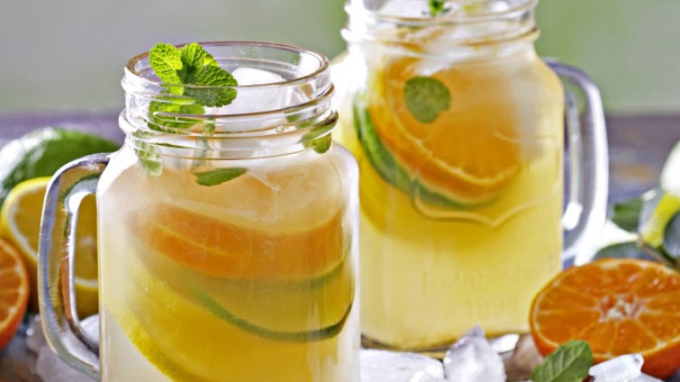 Zwei Gläser mit Henkel, gefüllt mit Eis, Limonade, Minzblättern und Scheiben von Orange, Limette und Zitrone, auf Eiswürfeln stehend.
