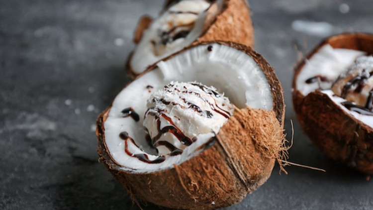 Halbe Kokosnüsse mit weißem Eis, dunkler Schokoladensoße und Kokosraspeln auf grauem Untergrund.