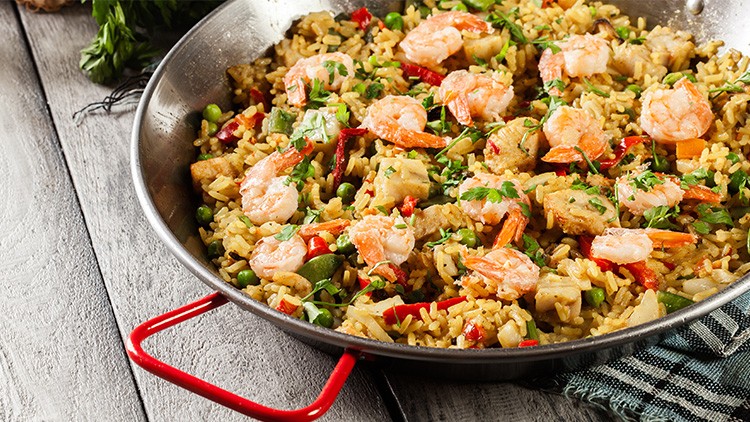 Eine silberne Pfanne mit Paella aus Reis, Garnelen, Paprika, Erbsen und Kräutern auf hellem Holztisch.