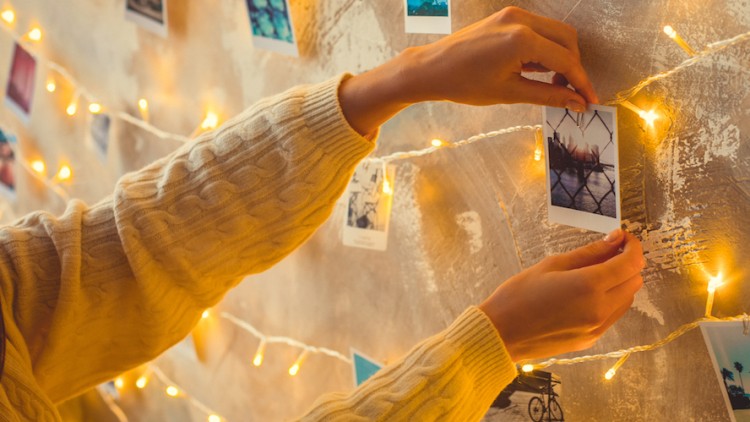 Hände in beigem Strickpullover befestigen ein Polaroidfoto an einer Lichterkette vor einer Wand mit weiteren Fotos und Lichtern.