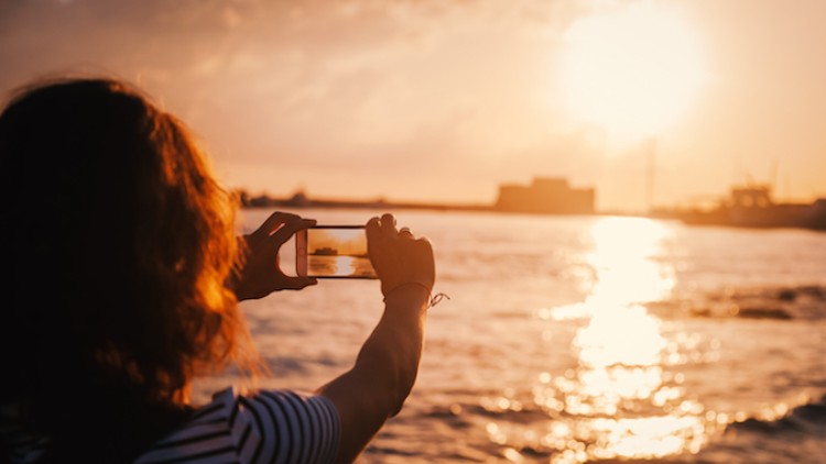 Person mit lockigen Haaren fotografiert einen Sonnenuntergang über einem Gewässer, die Sonne spiegelt sich im Wasser.