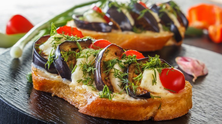 Getostete Brotscheiben mit gebratenen Auberginenscheiben, Kirschtomaten, frischen Kräutern und hellen Soßen auf dunklem Teller.