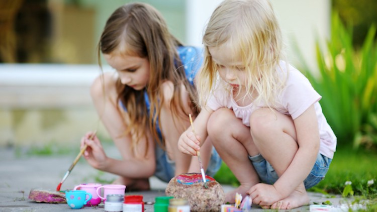 Zwei Kinder bemalen auf einer Terrasse einen Stein mit bunten Farben, um sie liegen Pinsel, Farbtöpfe und Spielzeug.