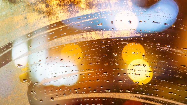 Wassertropfen auf einer beschlagenen Glasscheibe mit bunten, verschwommenen Lichtpunkten im Hintergrund.