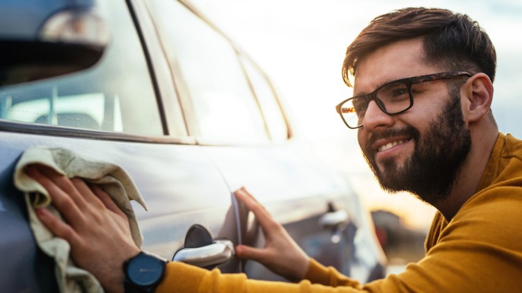 Eine Person reinigt mit einem Tuch die Autotür eines silbernen Autos bei Sonnenlicht.
