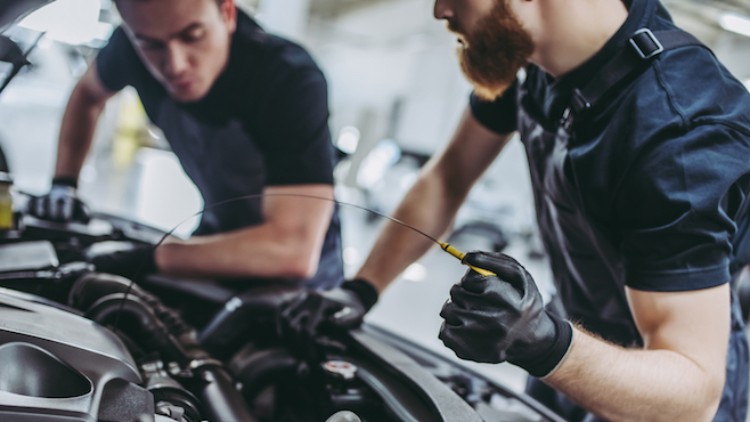 Zwei Personen in Arbeitskleidung überprüfen den Ölstand eines Autos in einer Werkstatt mit geöffnetem Motorraum.