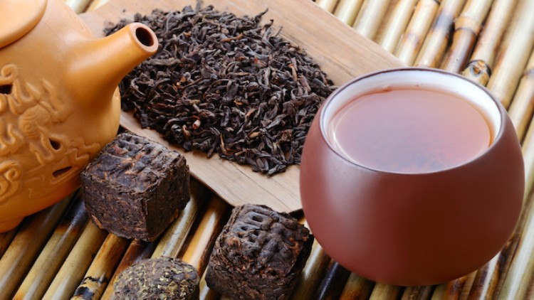 Braune Teetasse, tonfarbene Teekanne, Teeblätter auf Holzbrett und dunkle gepresste Teewürfel auf Bambusunterlage.