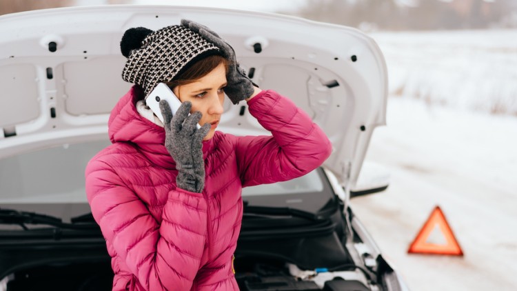Person in pinker Jacke telefoniert vor einem Auto mit geöffneter Motorhaube, im Hintergrund ein Warndreieck auf verschneiter Straße.