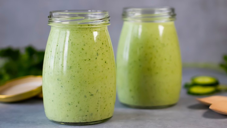 Zwei verschlossene Gläser mit grüner Sauce oder Dressing, daneben ein offener Deckel und unscharfes Gemüse im Hintergrund.