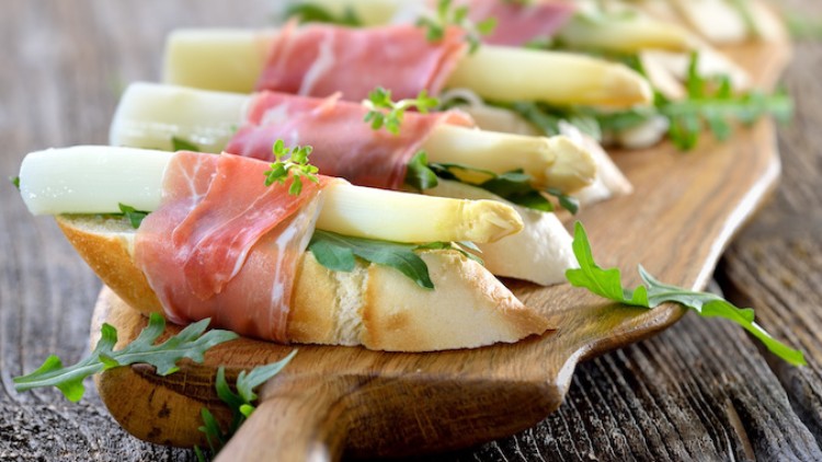Scheiben Baguette mit weißem Spargel, Schinken, Rucola und Kräutern auf einem hölzernen Servierbrett.