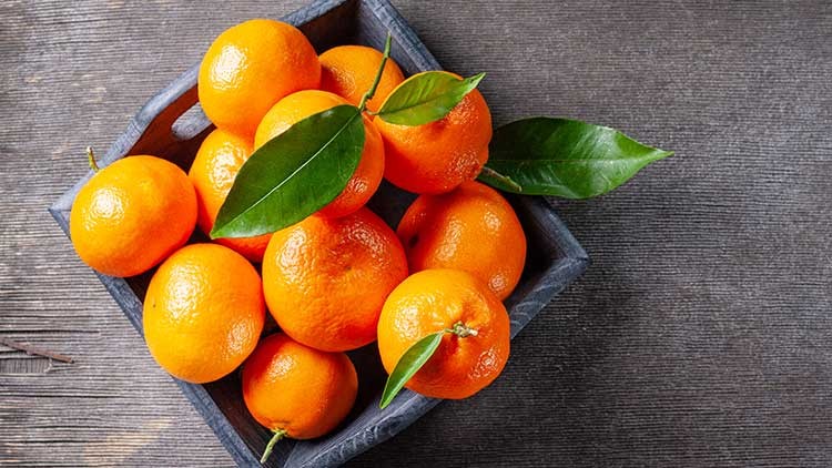 Mehrere leuchtend orange Mandarinen mit grünen Blättern liegen in einer dunklen Holzschale auf einem Holztisch.