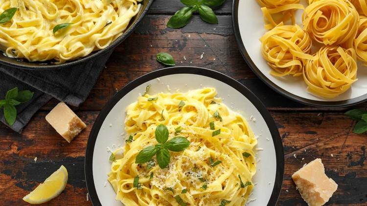 Ein Teller Tagliatelle mit heller Sauce, Parmesan und Basilikum, daneben Tagliatelle-Nester auf einem Teller und ein Parmesanstück.