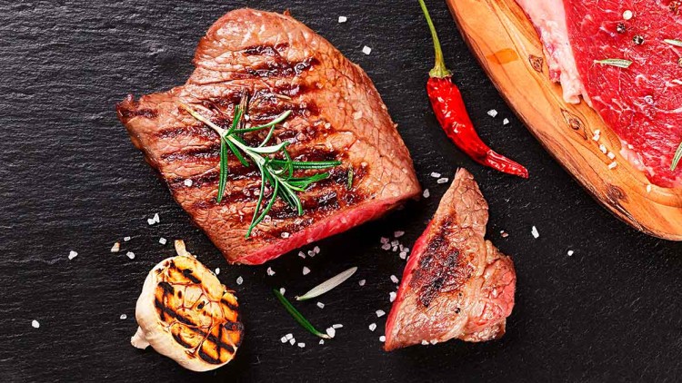 Gegrilltes Steak mit Rosmarin, gerösteter Knoblauch, rohes Fleisch auf Holzbrett, rote Chili und grobes Salz auf schwarzem Untergrund.