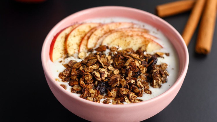 Rosa Schüssel mit Joghurt, Müsli, frischen Apfelscheiben und Zimt auf schwarzem Untergrund, daneben zwei Zimtstangen.