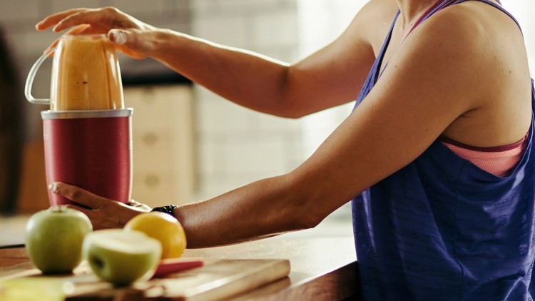 Person in Sportkleidung steckt Zutaten in einen roten Mixer, daneben frisches Obst und ein Glas Orangensaft auf einem Holztisch.